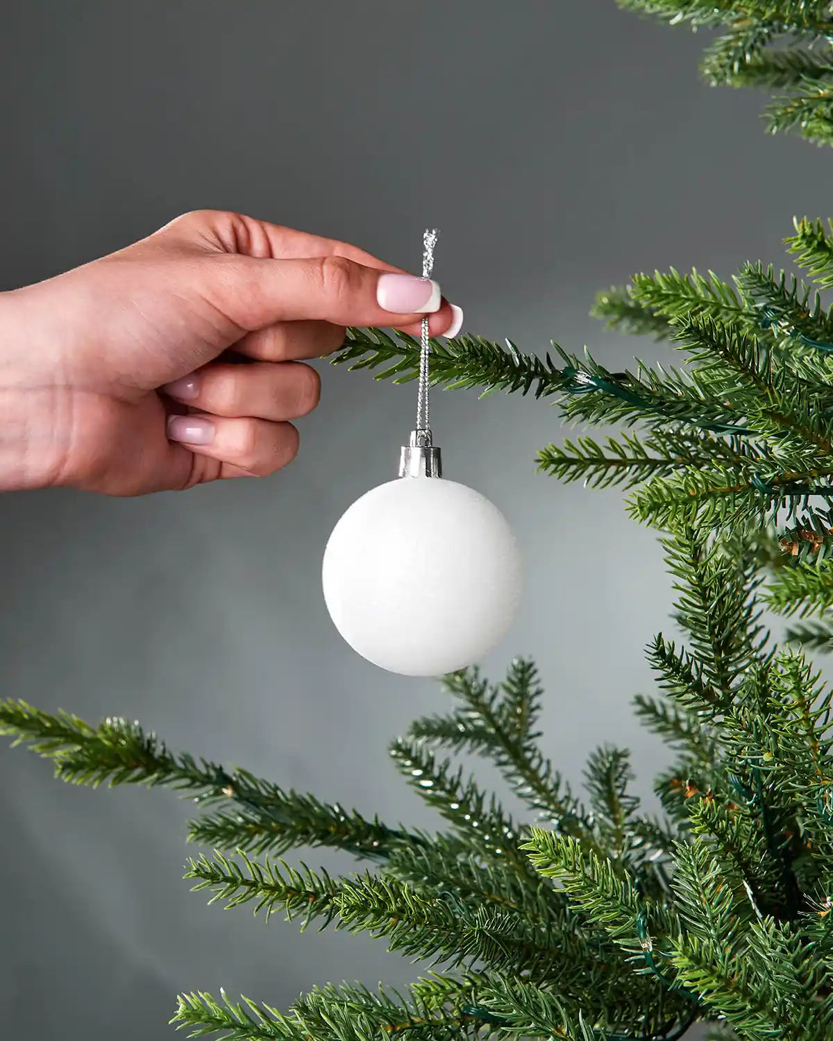 Hand placing a white Christmas bauble on a tree branch against a grey background