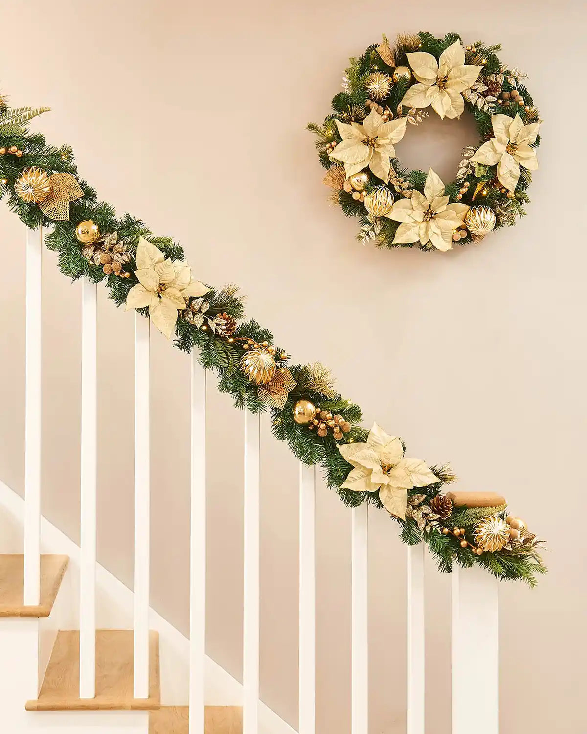 Decorative Christmas garland and matching wreath with gold poinsettias, pinecones, and lights on a staircase banister neutral background.