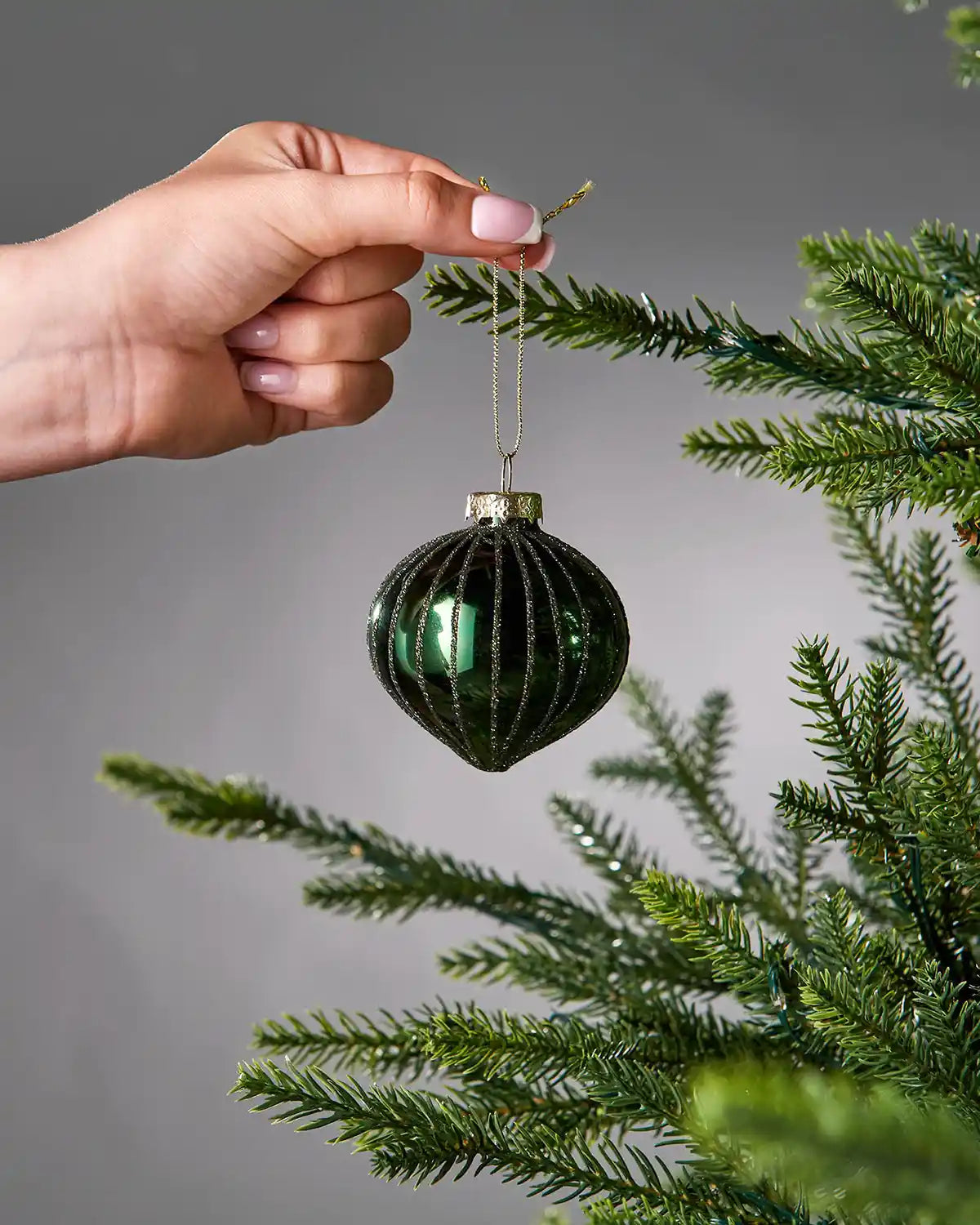 Hand placing a green glass bauble on a Christmas tree branch against a grey background