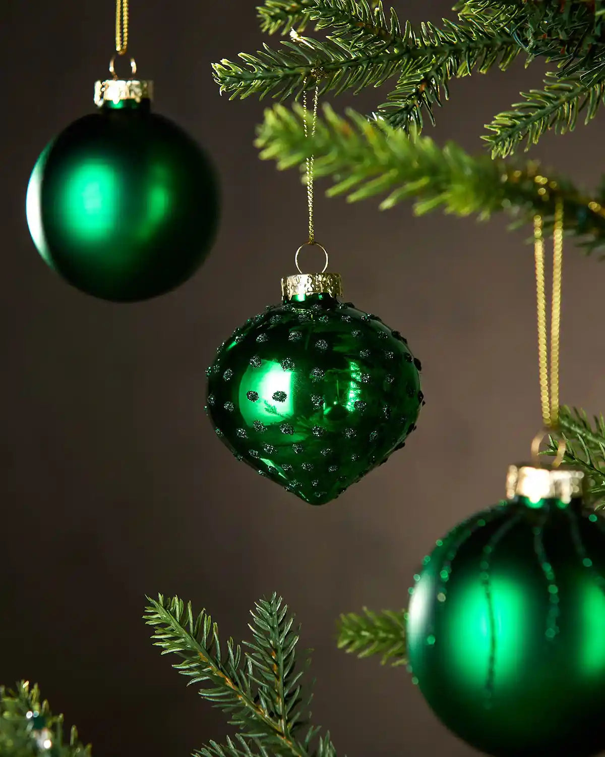 Three Green Christmas baubles with glitter and textured hanging on a tree branch against a dark background