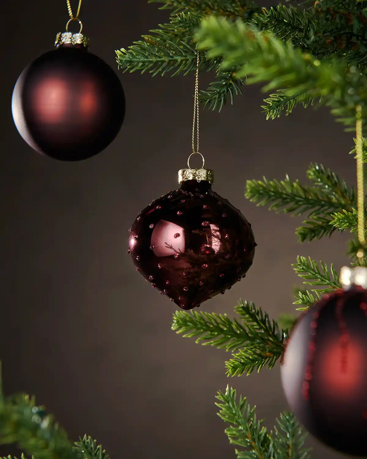 Three Burgundy glass ornaments hanging on a Christmas tree with a dark background