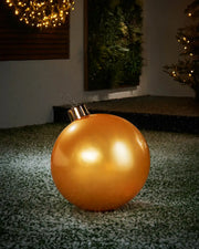 Gold Medium inflatable bauble on snowy grass with a decorated tree and wreath in background