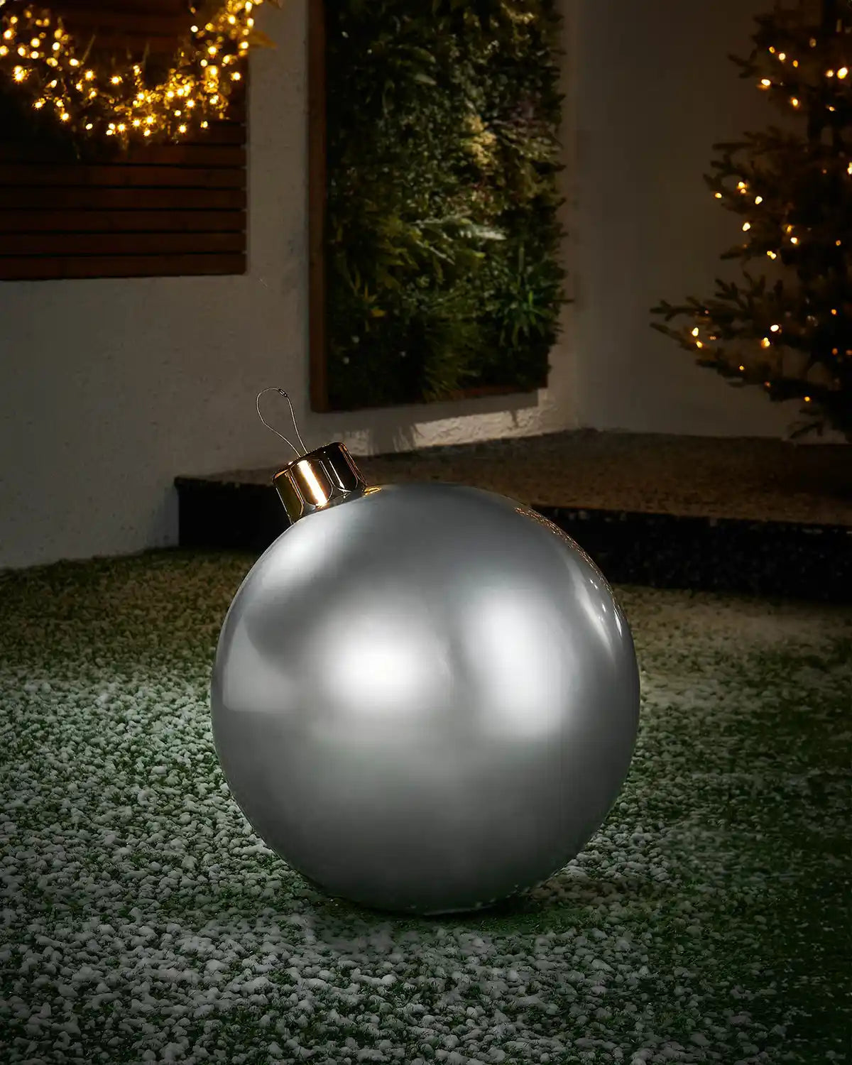 Medium sized silver inflatable bauble on a snowy garden floor with a decorated Christmas tree and wreath in background 
