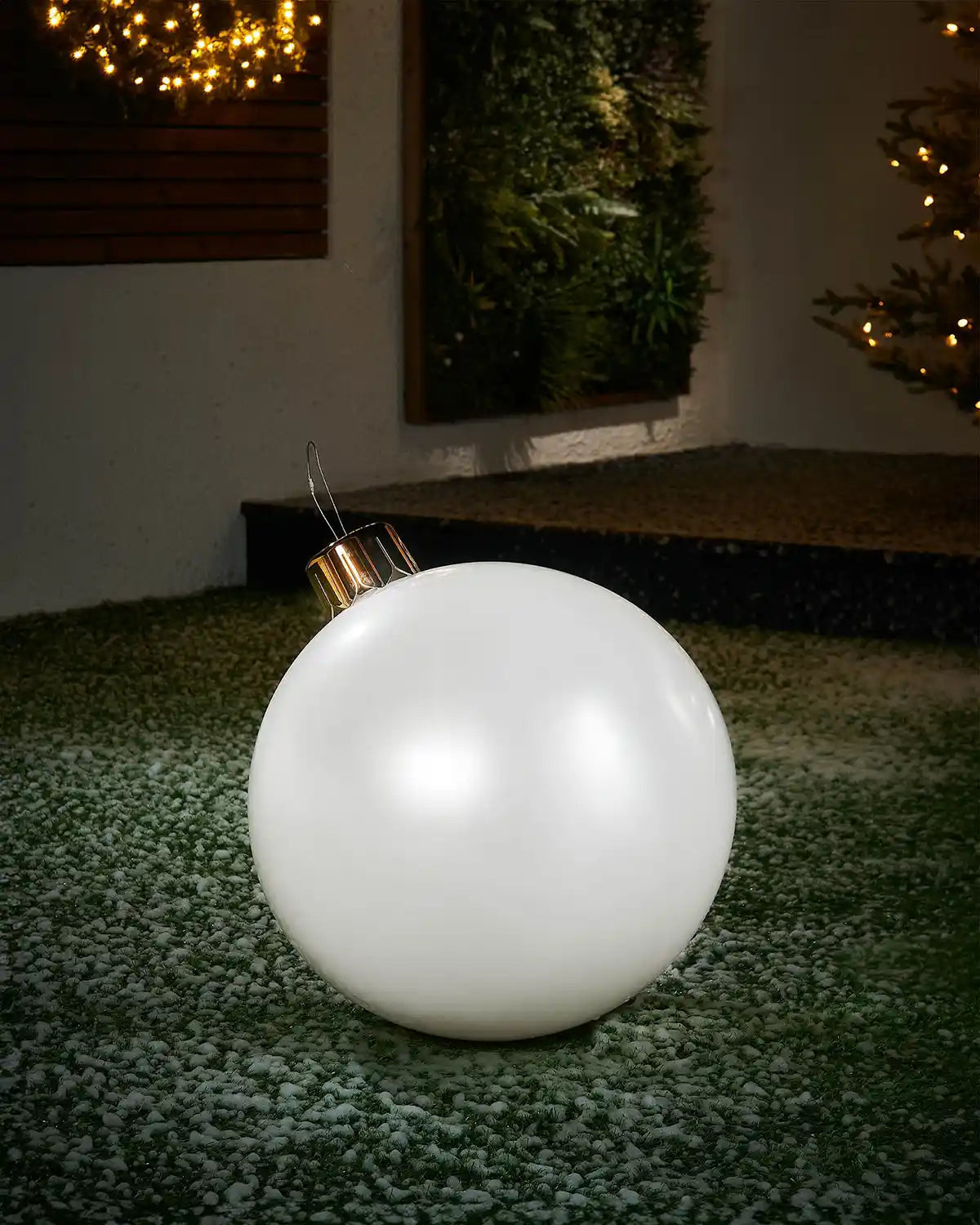 Medium sized White inflatable bauble on a snowy garden floor with a decorated Christmas tree and wreath in background 
