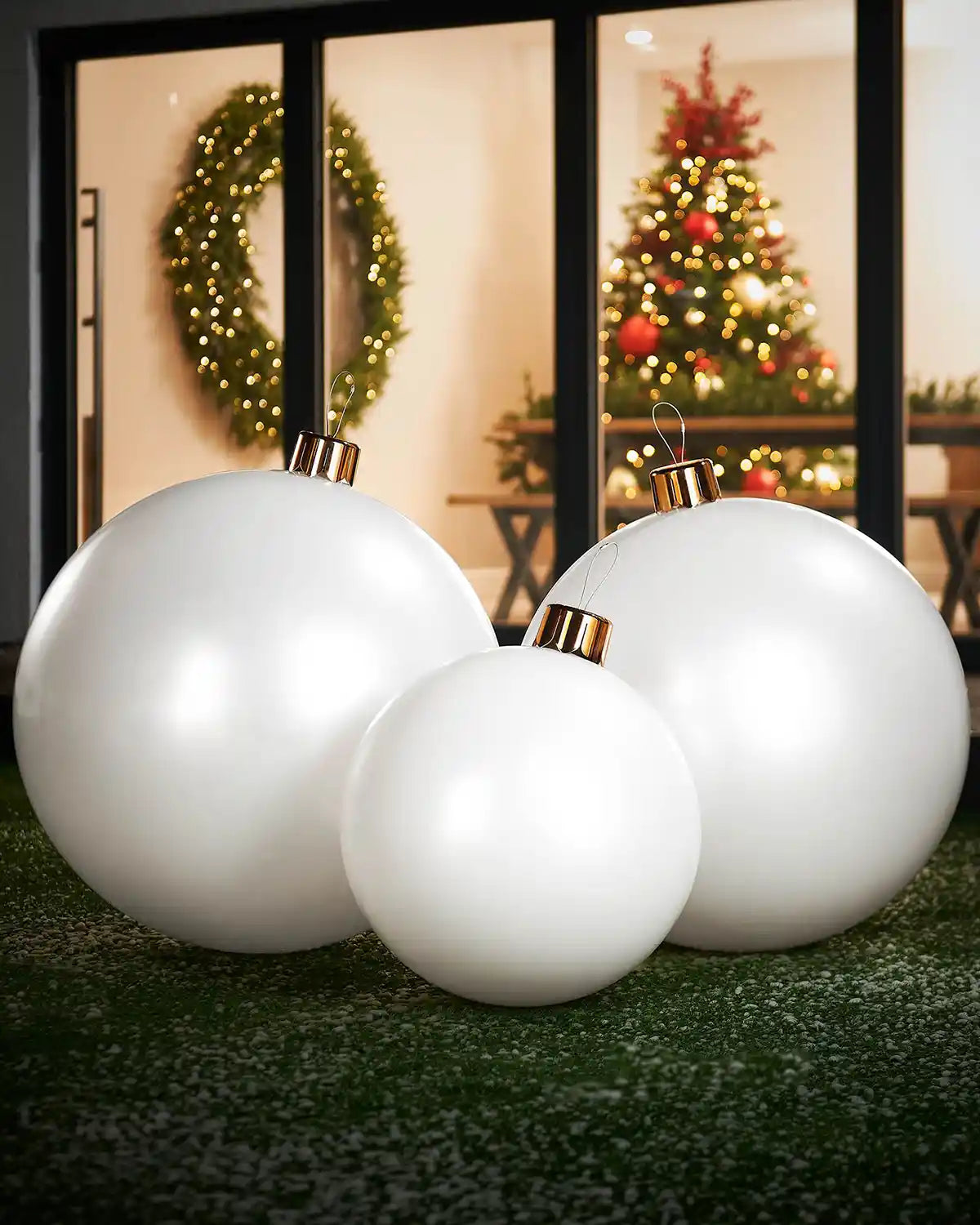 Medium, large and extra large white Christmas ornaments on a grassy surface with a festive indoor background.

