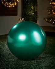 Large sized green inflatable bauble on a snowy garden floor with a decorated Christmas tree and wreath in background 
