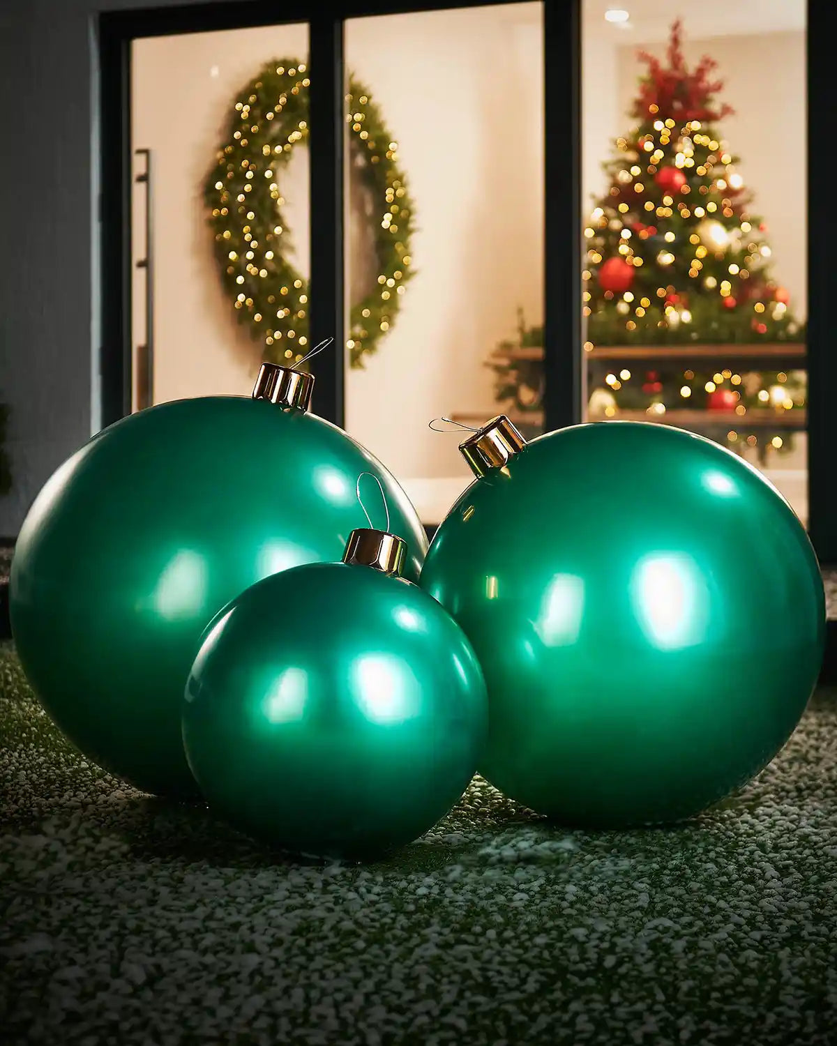Medium, large and extra large green Christmas inflatable baubles on a grassy surface with a festive indoor background.
