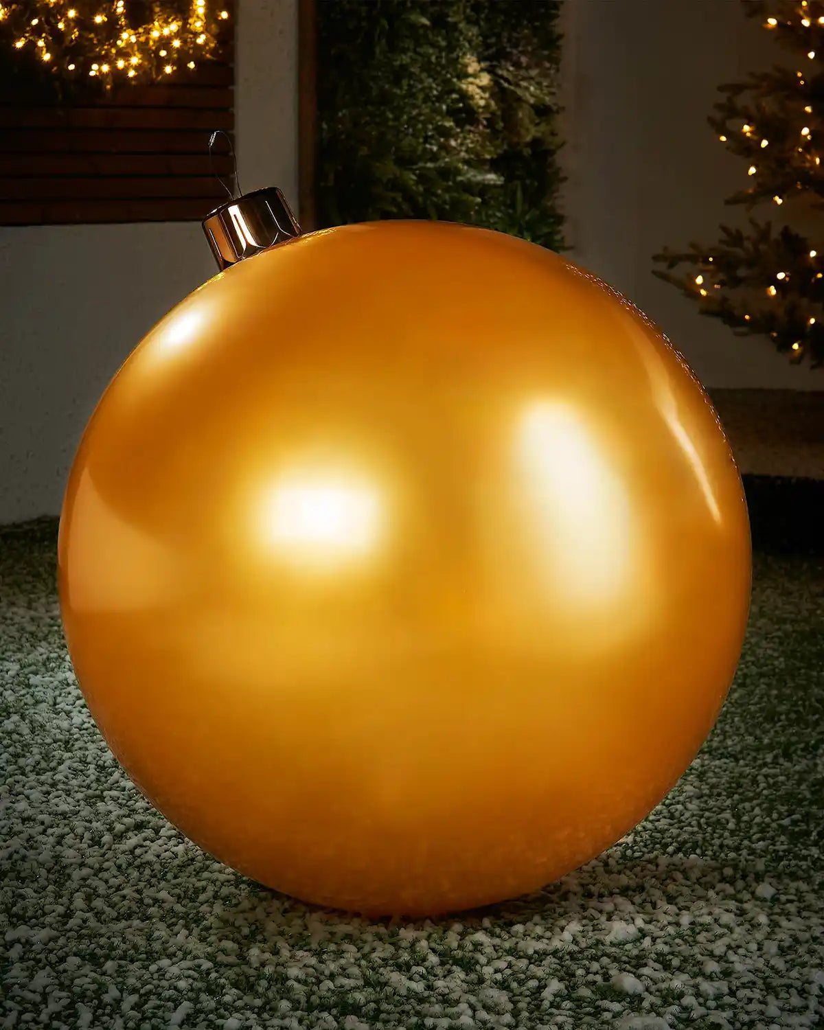Extra large sized gold inflatable bauble on a snowy garden floor with a decorated Christmas tree and wreath in background 
