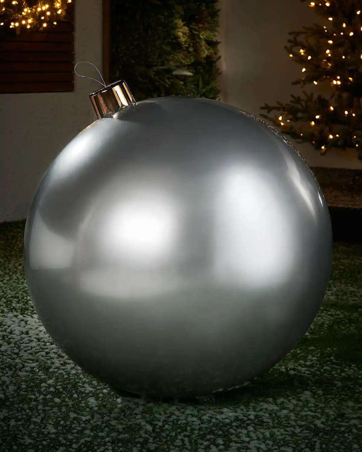 Extra large sized silver inflatable bauble on a snowy garden floor with a decorated Christmas tree and wreath in background 
