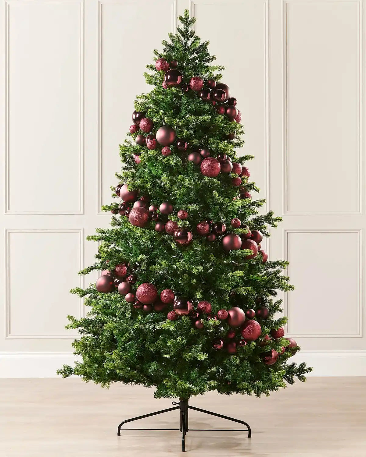 Burgundy bauble garland wrapped around a Christmas tree against a cream panel wall and a wooden floor 

