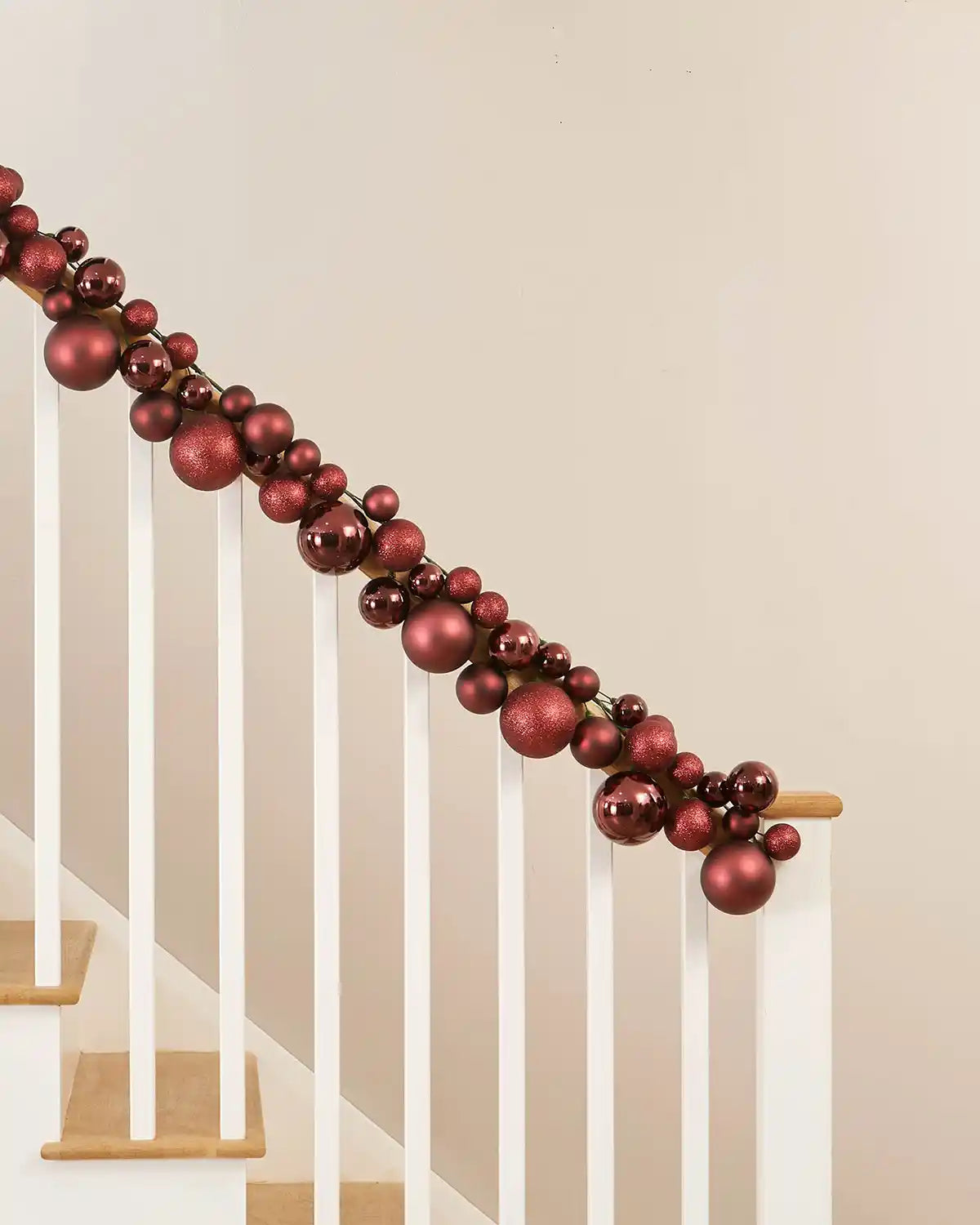 Decorative burgundy bauble garland on a staircase railing against a beige wall.
