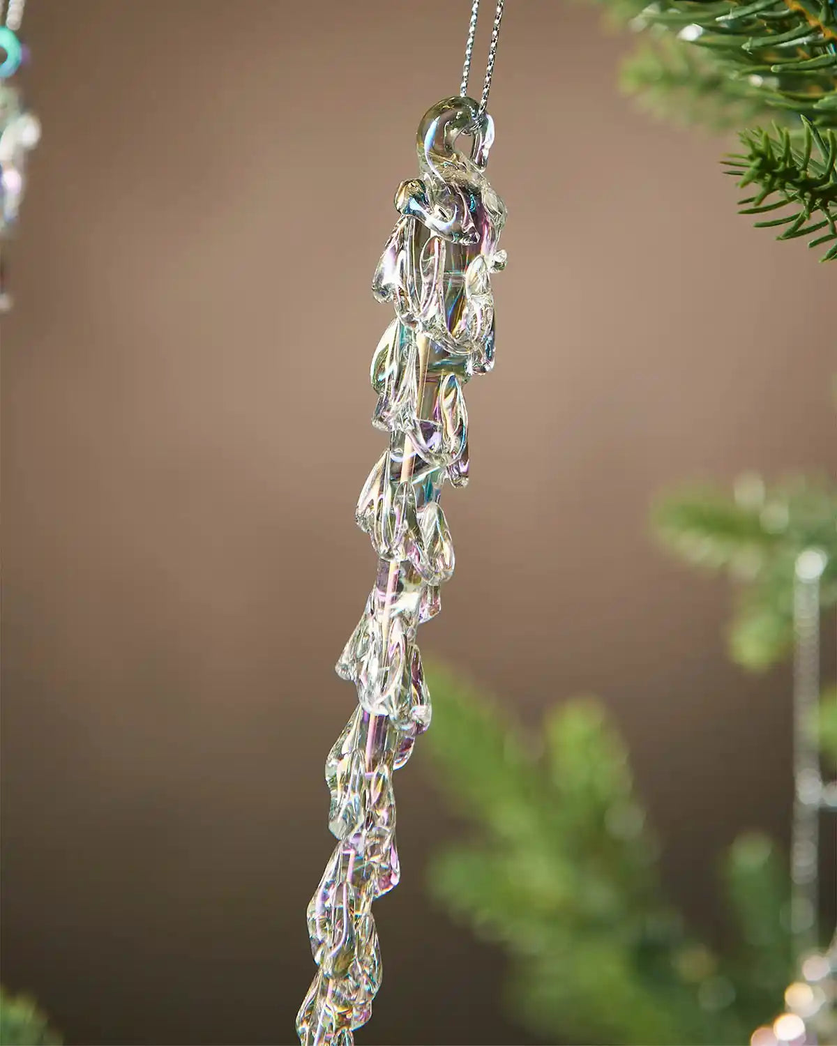 Close up of an iridescent crystal icicle ornaments hanging from a Christmas tree branch against a brown background  