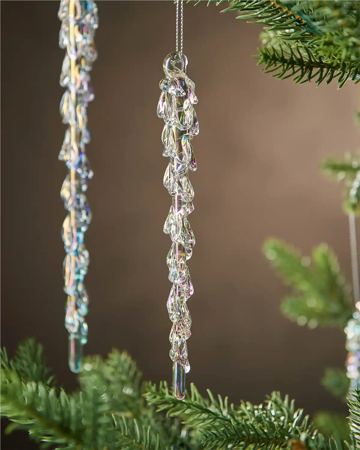 Close up of two iridescent crystal icicle ornaments hanging from a Christmas tree branch against a brown background 