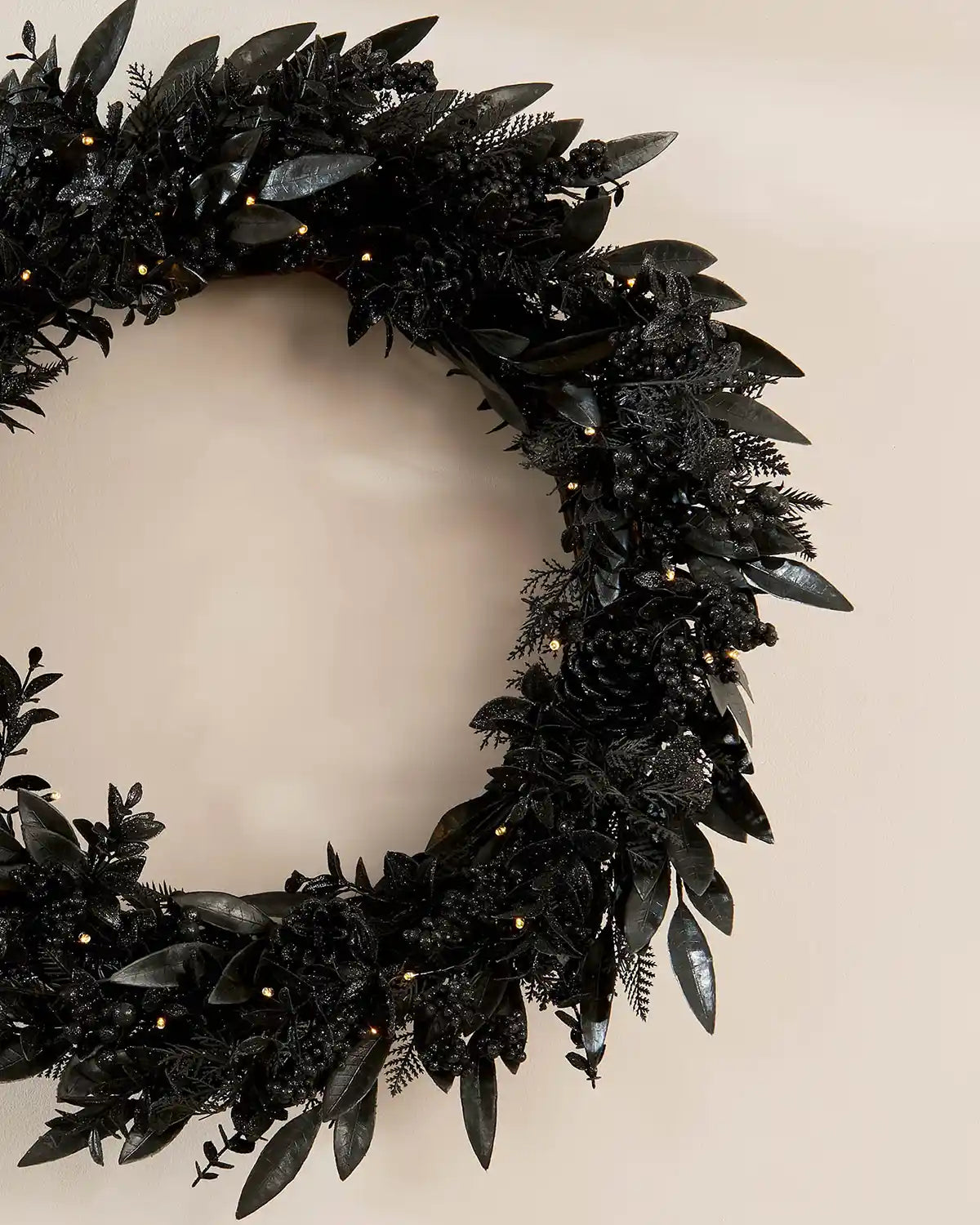 Close up of Black decorated wreath with black pine cones, berries and warm white lights against a cream wall
