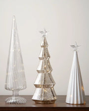 Set of three glass shaped Christmas trees with warm white lights against a white wall on a wooden table 