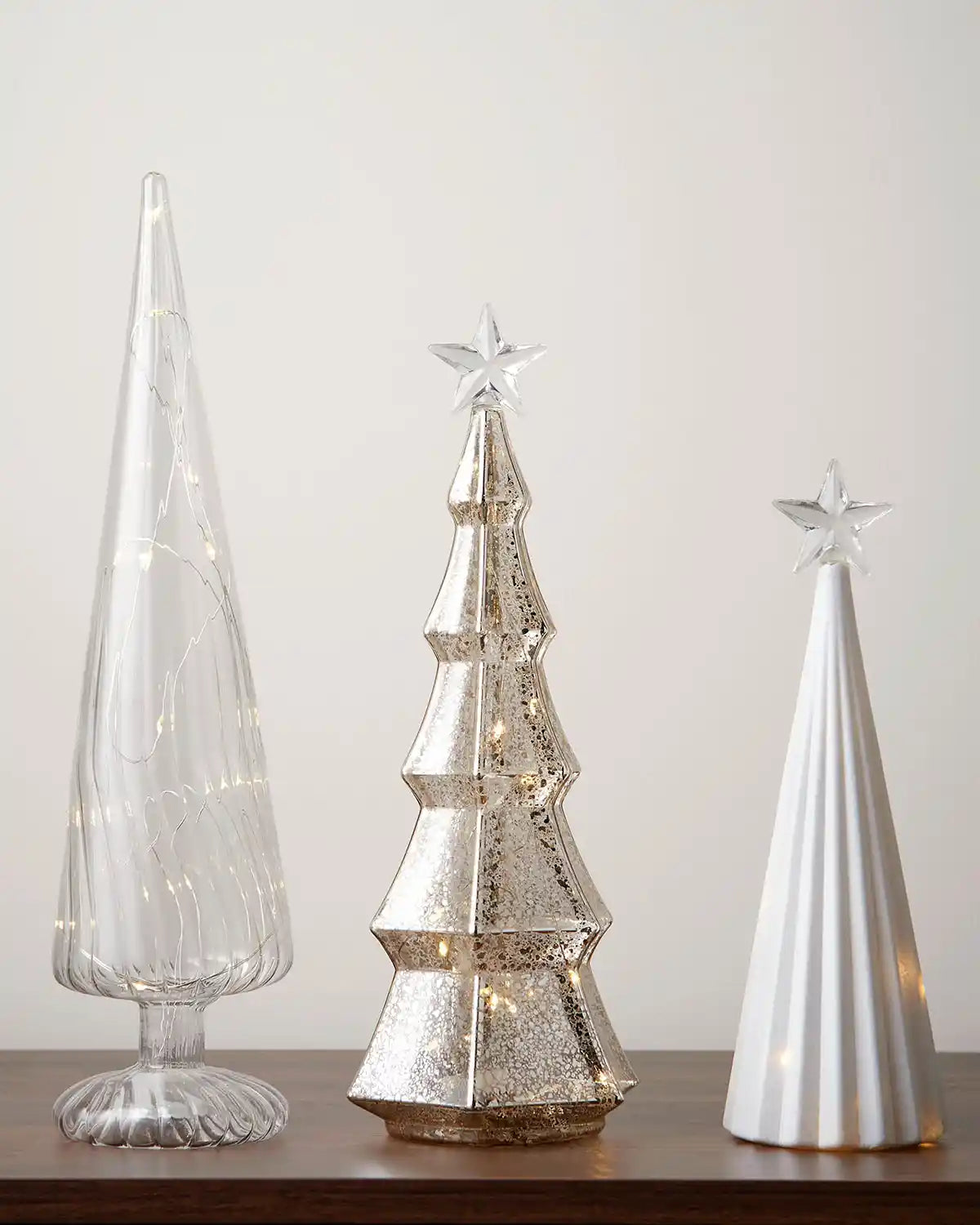 Set of three glass shaped Christmas trees with warm white lights against a white wall on a wooden table 