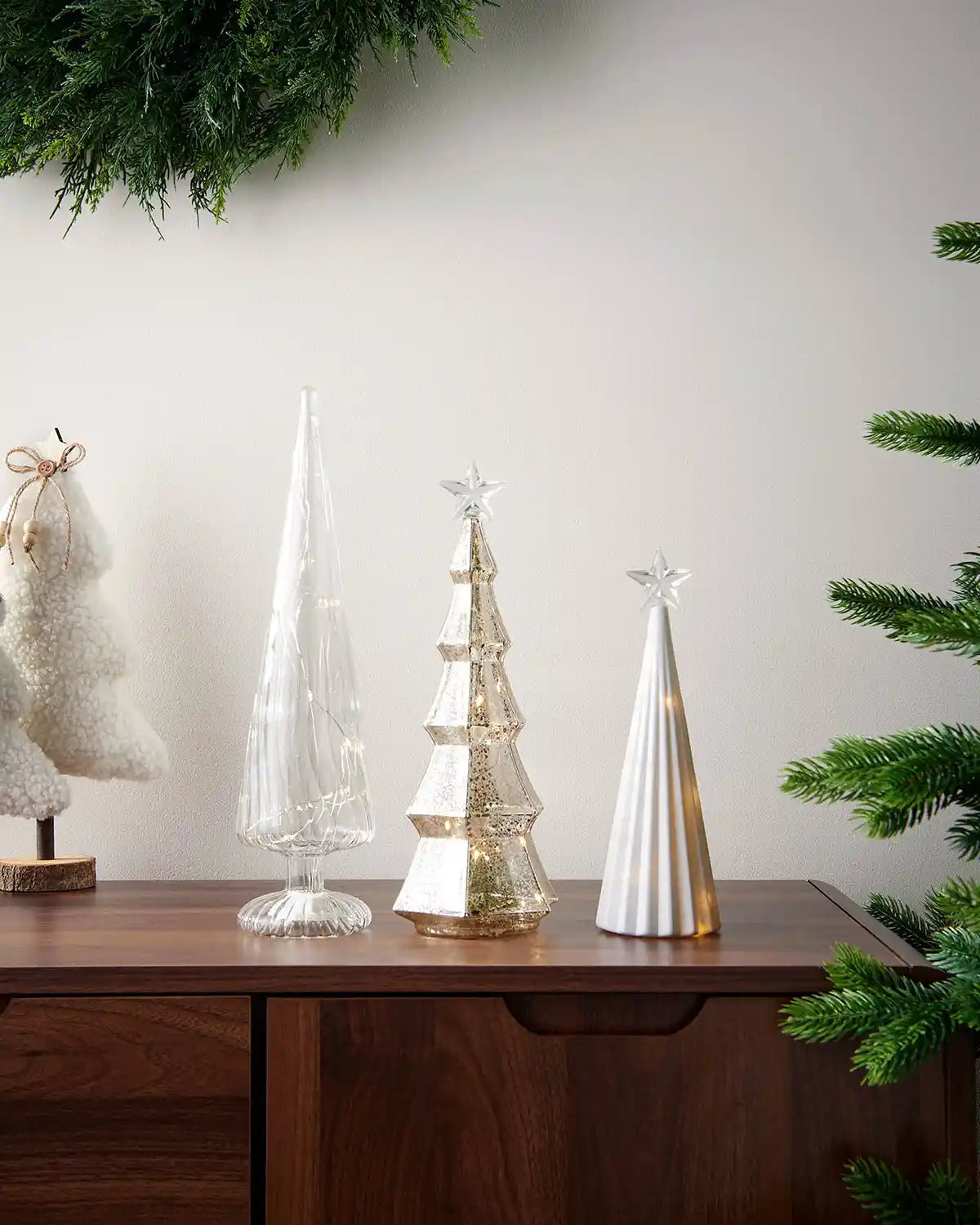 Set of three glass Christmas trees with warm white lights on a wooden table and a Christmas tree in background