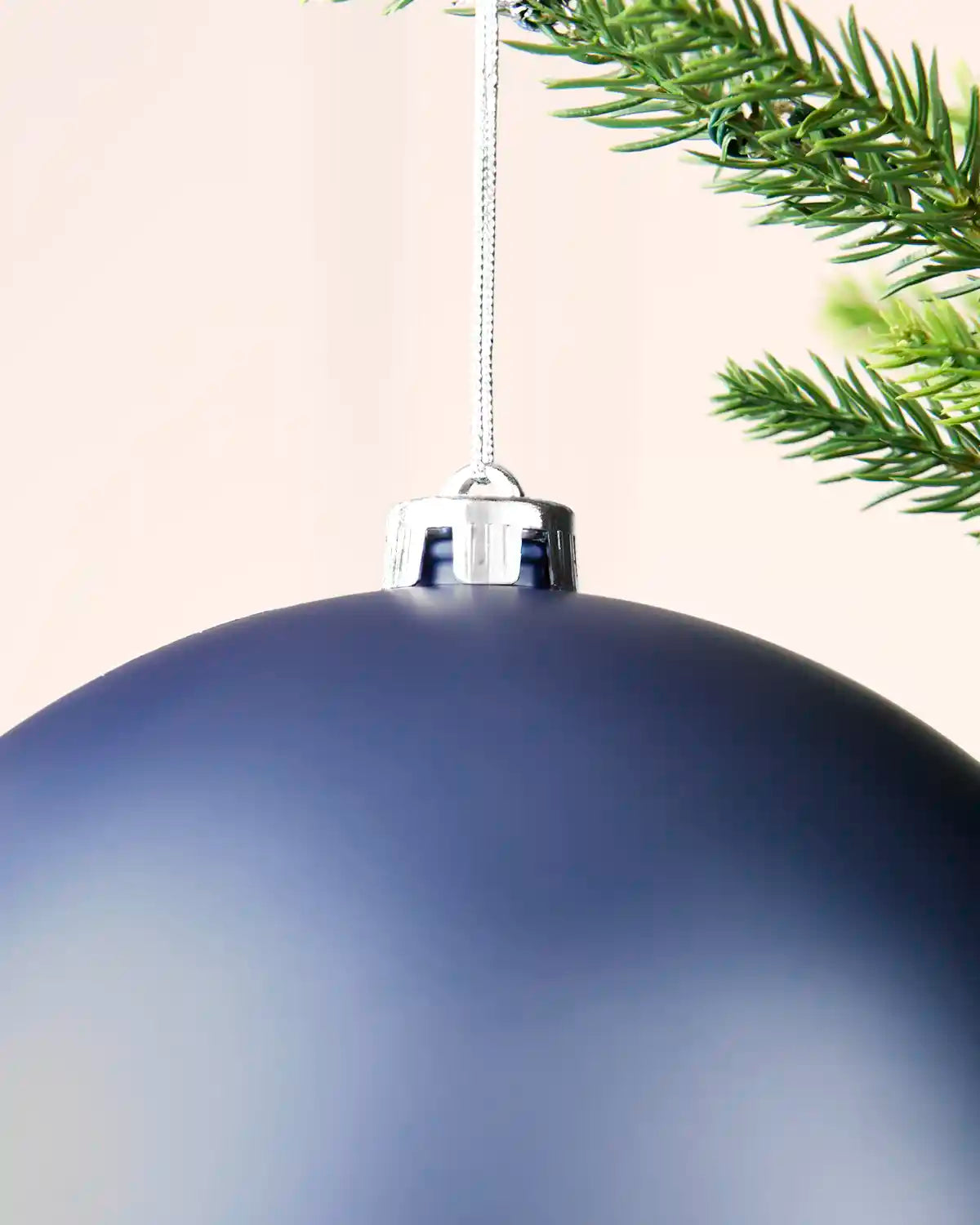 Close up of a large matte blue Christmas bauble hanging from a silver cap on a tree branch, with green pine needles and part of another ornament visible.