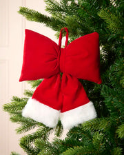 A red and white faux fur Santa hat Christmas ornament shaped like a large bow, hanging on a green Christmas tree.