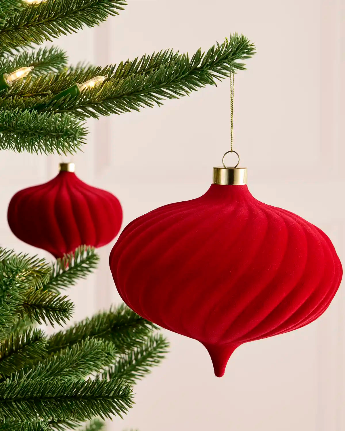 A red ribbed Christmas ornament hanging from a green Christmas tree branch, with a second matching ornament in the background against a soft light beige wall.