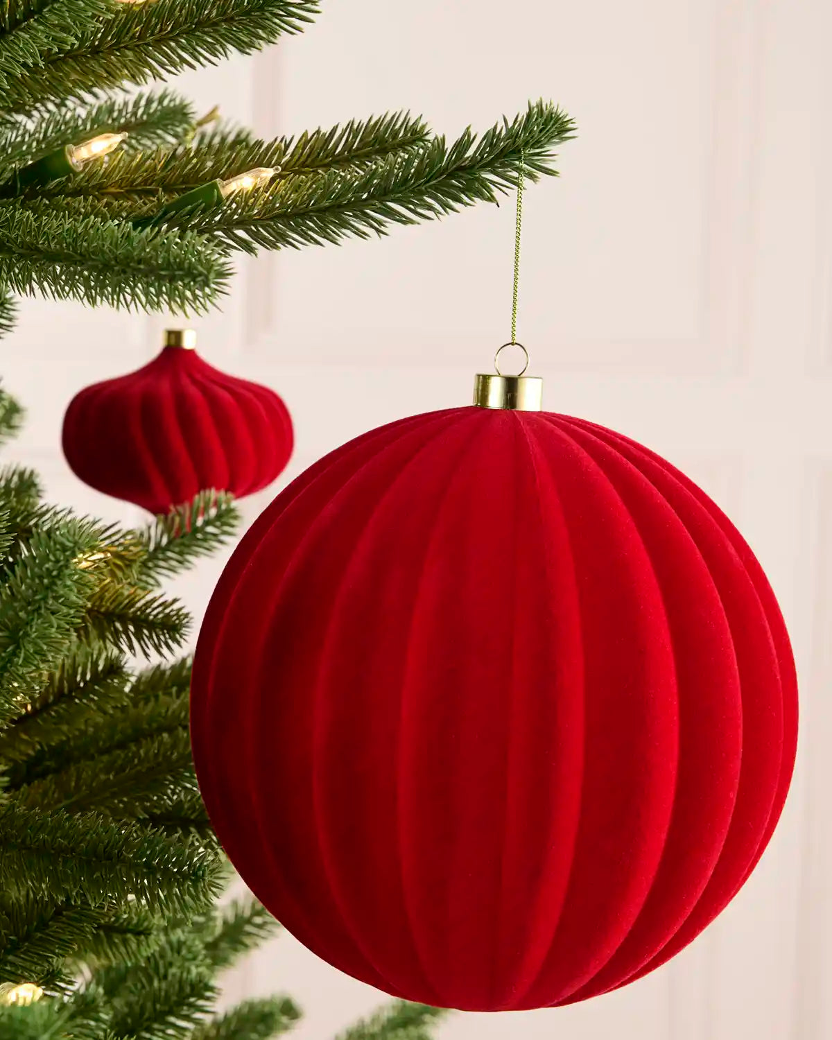 A large red ribbed velvet Christmas ornament hanging from a green Christmas tree branch, with a smaller matching ornament blurred in the background.