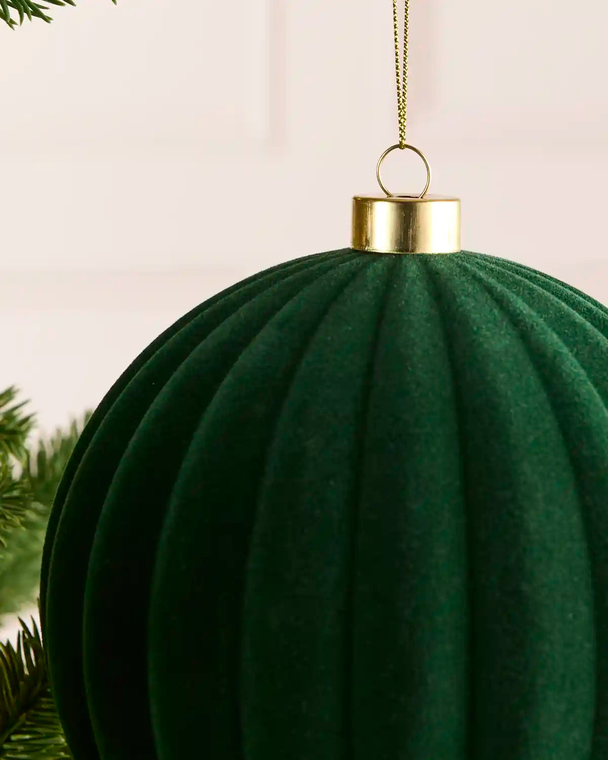 A close-up of a sage green ribbed fabric Christmas ornament hanging from a gold cap and chain, with a soft neutral background and a hint of pine branches at the edge.