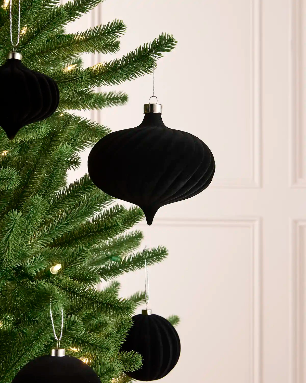 Black ribbed velvet Christmas tree ornaments hanging on a green pine tree with warm white string lights against a light cream paneled wall.