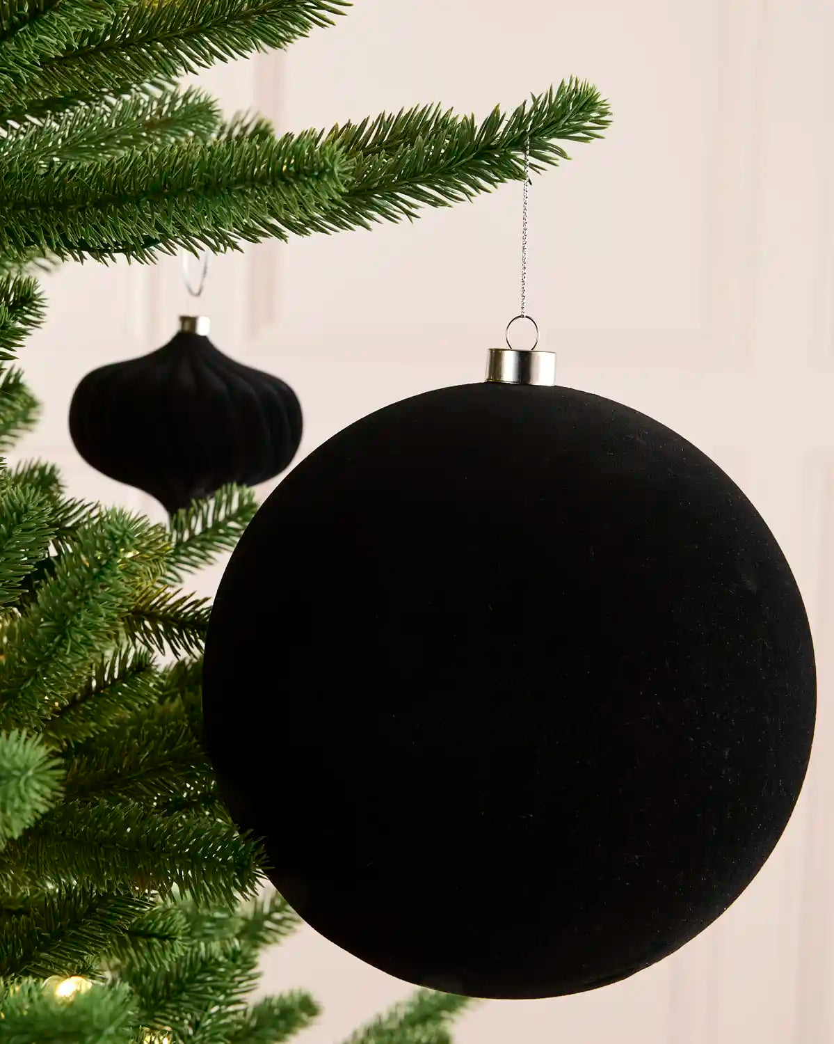A large black velvet Christmas bauble hanging from a green pine tree branch, with a smaller matching black ornament in the background against a soft light pink wall.