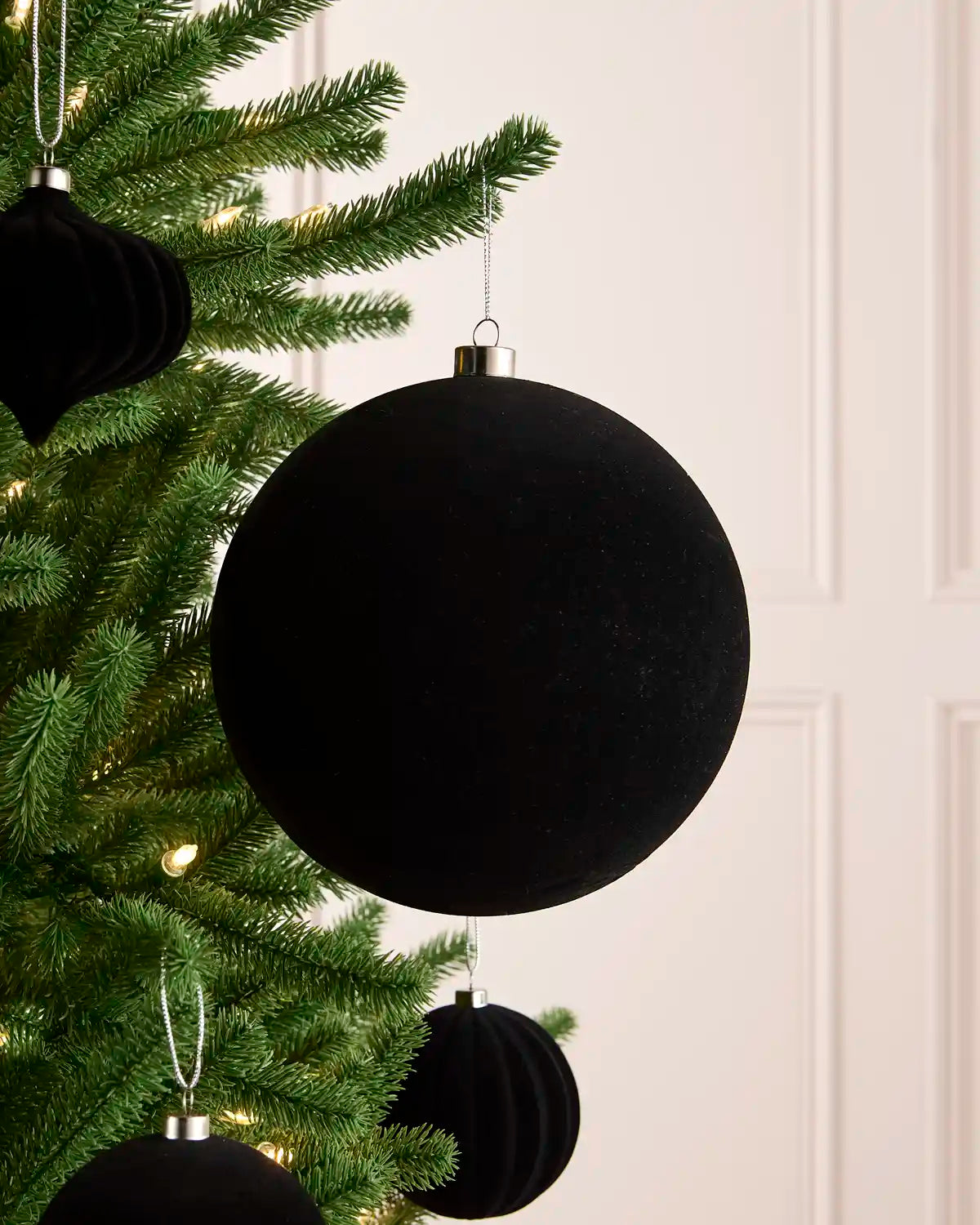 A large black velvet Christmas bauble hanging from a green pine tree branch, with smaller matching black ornaments in the background against a light wall.