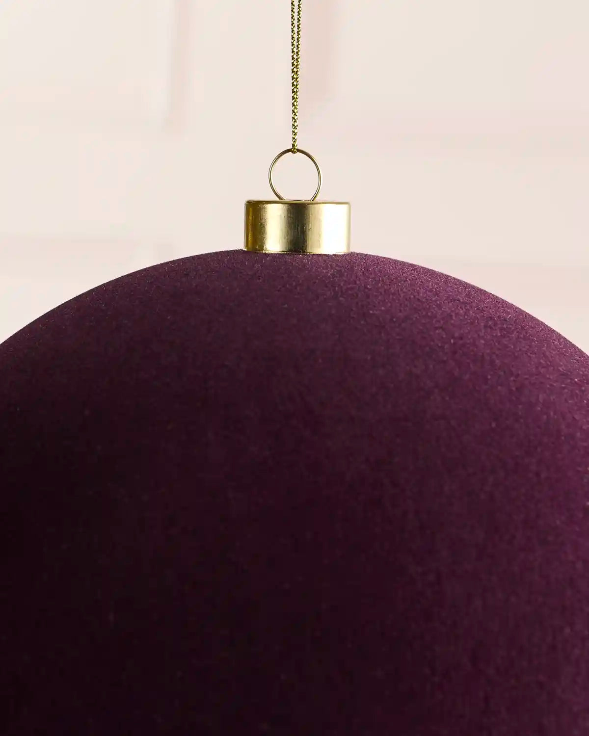 A close-up of a large, velvet purple Christmas ornament with a gold metal cap and hanging loop, suspended by a thin gold string against a soft, light beige background.
