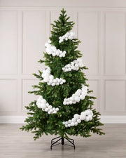 A green artificial Christmas tree with white bubble ornaments, displayed on a black metal stand against a light beige paneled wall and wood floor.