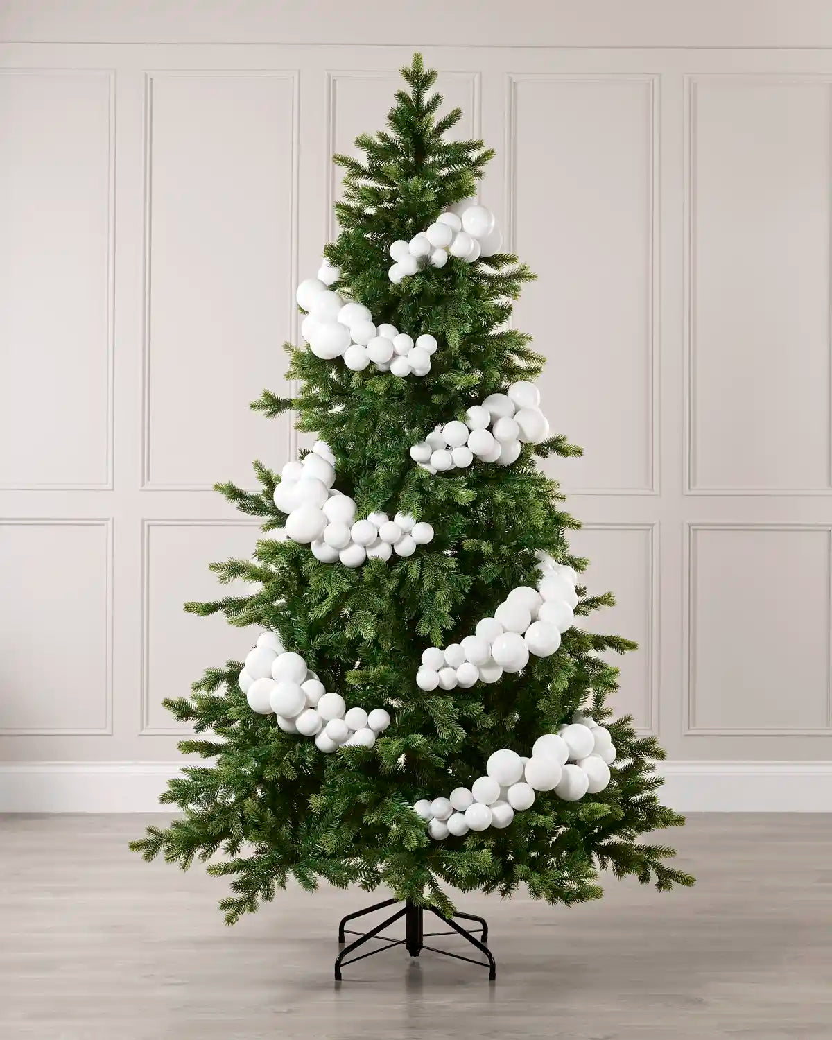 A green artificial Christmas tree with white bubble ornaments, displayed on a black metal stand against a light beige paneled wall and wood floor.