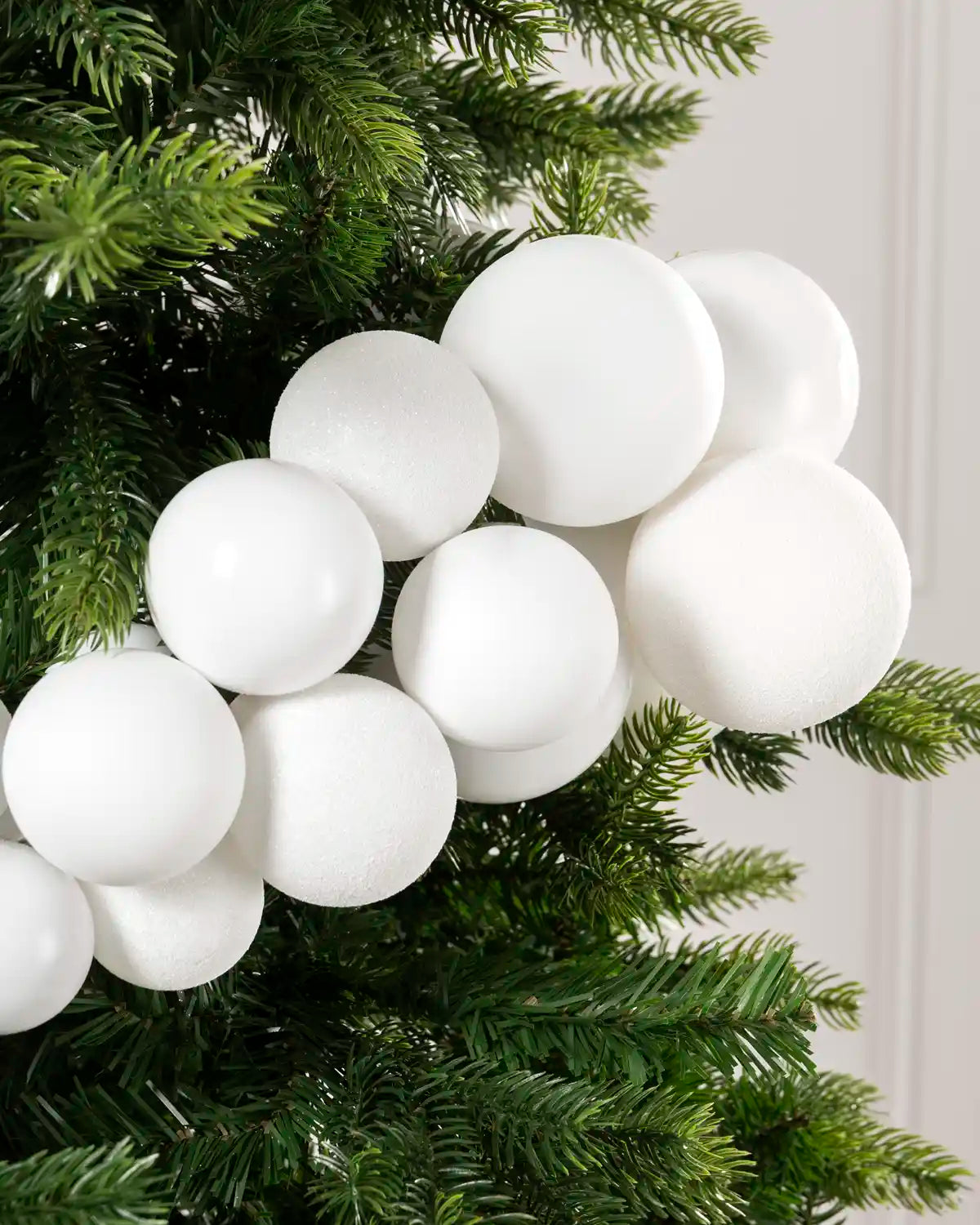 A cluster of matte white spherical ornaments hanging from the green branches of a Christmas tree against a light neutral wall.