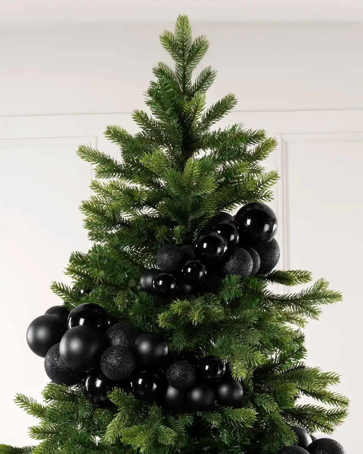 A green artificial Christmas tree decorated with matte black and textured black spherical ornaments, shown against a plain white wall.