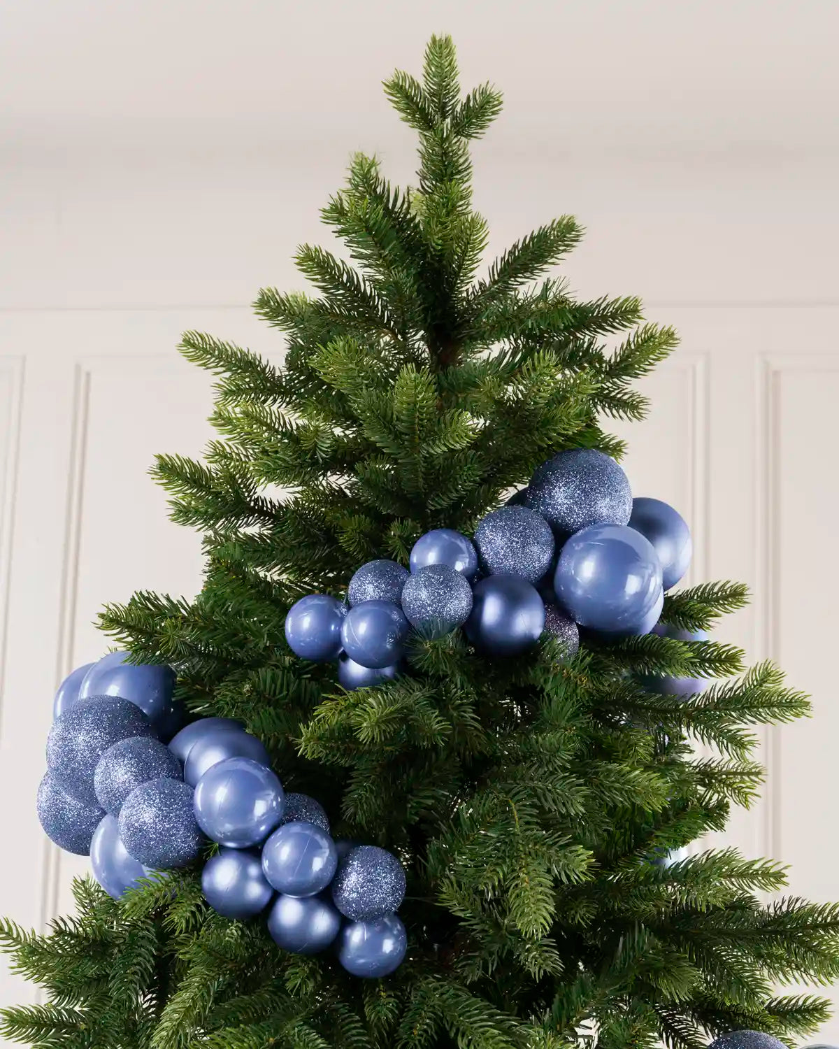 Ice Blue decorative ornaments, including shiny and glittery round baubles, arranged on a green artificial Christmas tree against a light paneled wall.