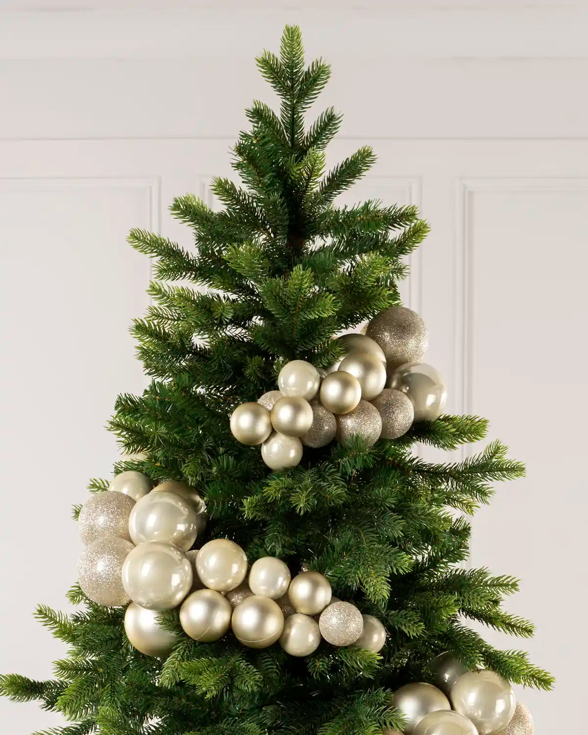 A green artificial Christmas tree decorated with clusters of metallic Latte t baubles, set against a light neutral paneled wall.