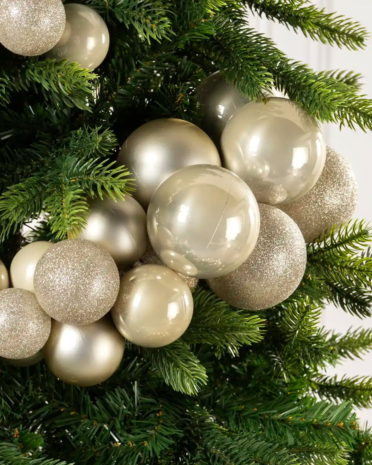 A close-up of round Christmas tree ornaments in metallic Latte, glittery, and matte, hanging on green artificial pine branches.