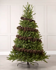 A green artificial Christmas tree decorated with multiple Espresso coloured spherical ornaments, standing on a black metal stand against a white paneled wall and light gray floor.