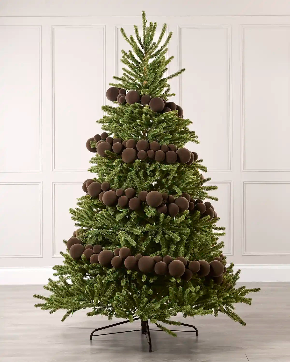 A green artificial Christmas tree decorated with multiple Espresso coloured spherical ornaments, standing on a black metal stand against a white paneled wall and light gray floor.