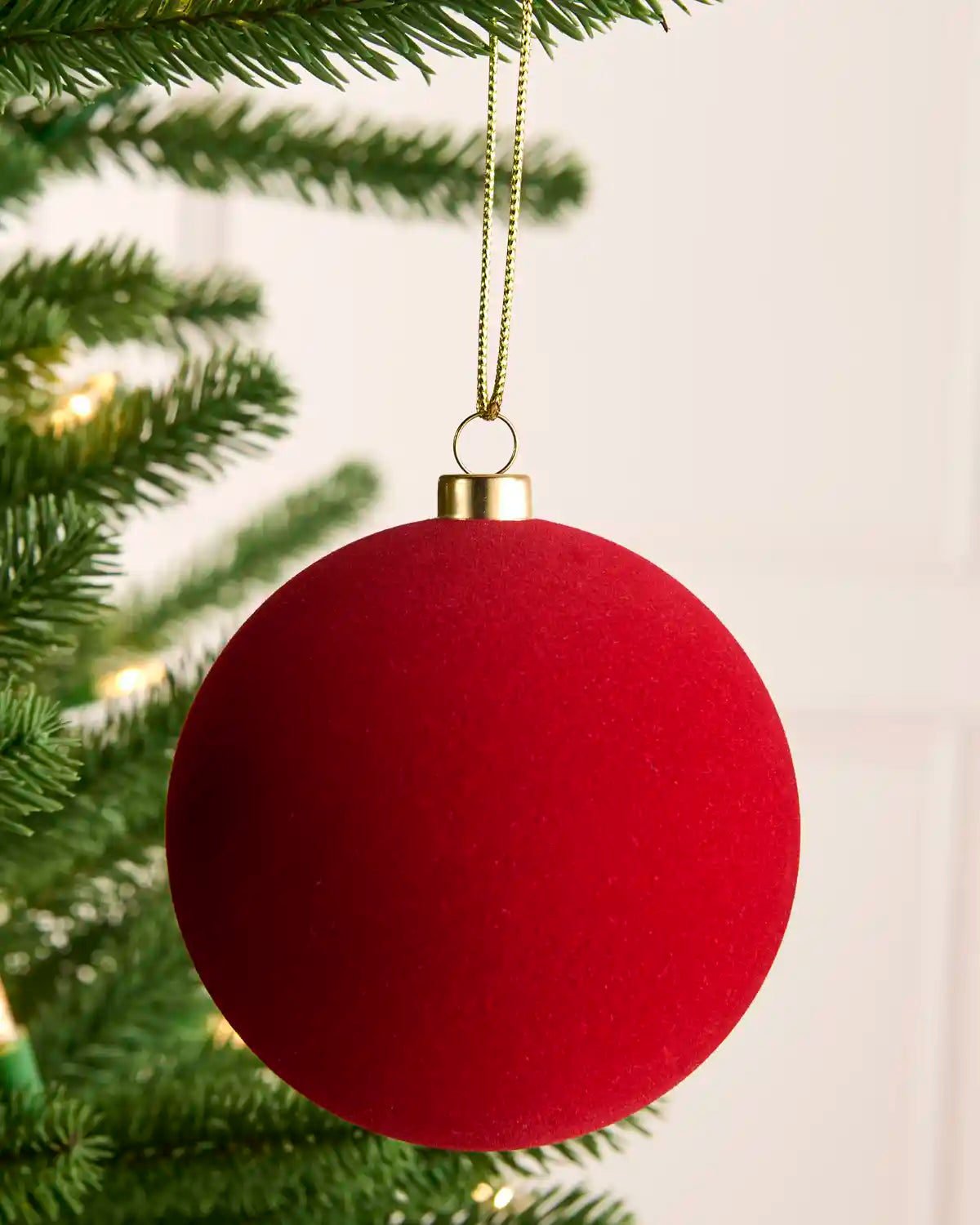 A red smooth velvet bauble hanging on a Christmas tree with gold string against a light background