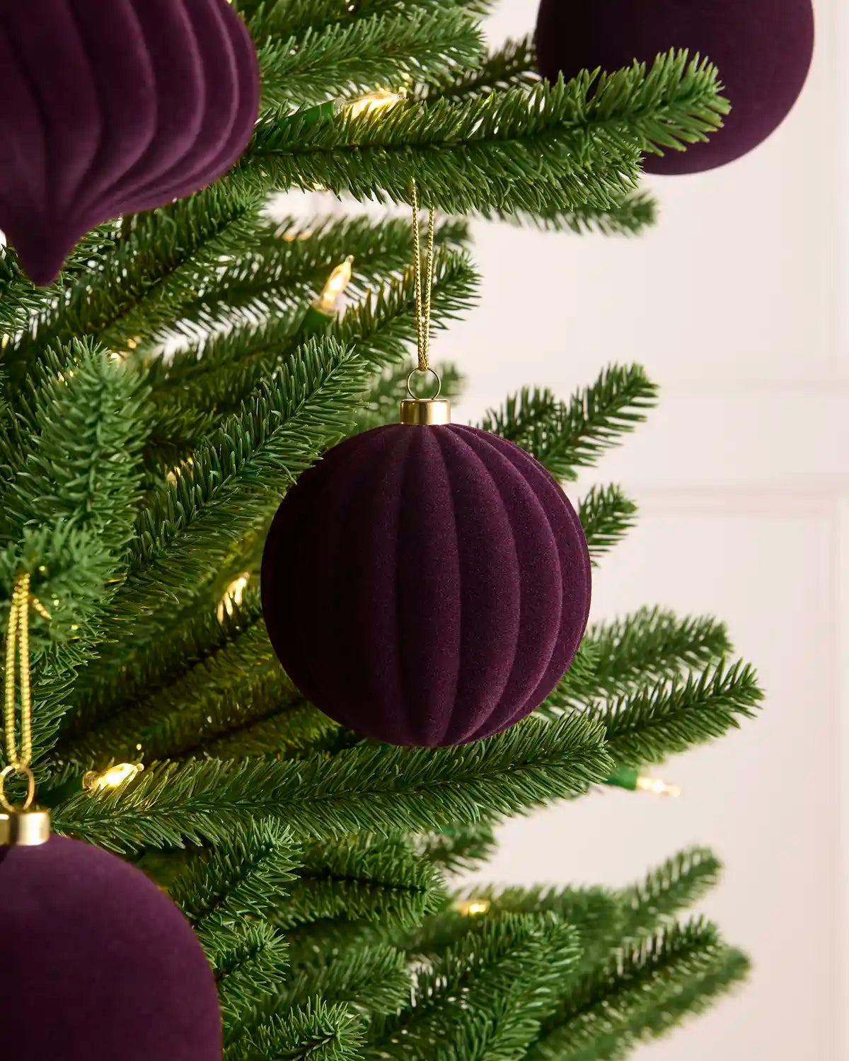 A close-up of a green Christmas tree decorated with ribbed, purple velvet ball ornaments and small warm white string lights against a soft off-white background.