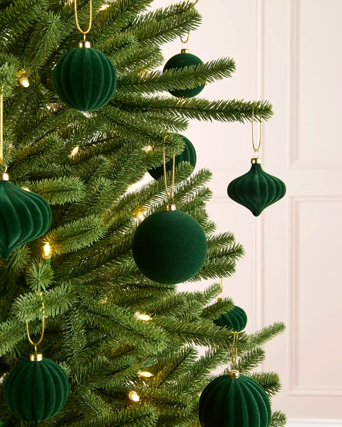 Sage green velvet Christmas tree ornaments hanging on a green artificial Christmas tree with warm white string lights against a light panelled wall.