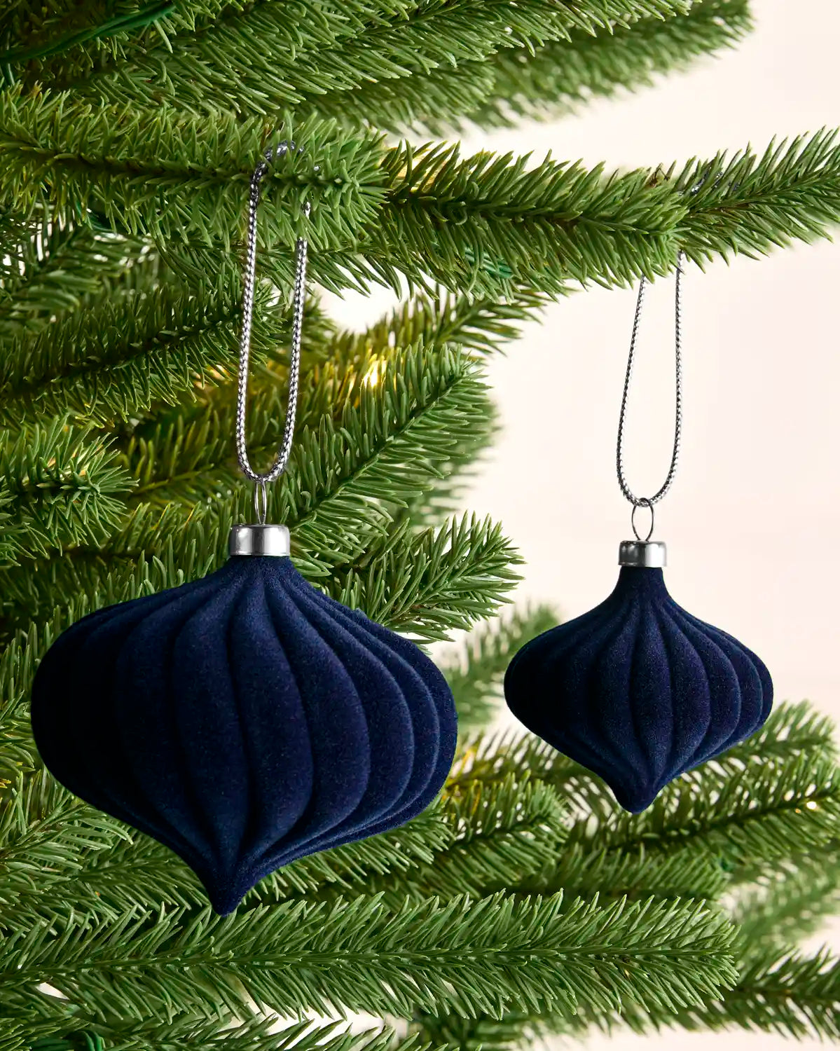 Two dark navy blue ribbed velvet Christmas tree ornaments hanging from silver chains on green pine tree branches against a light cream background.