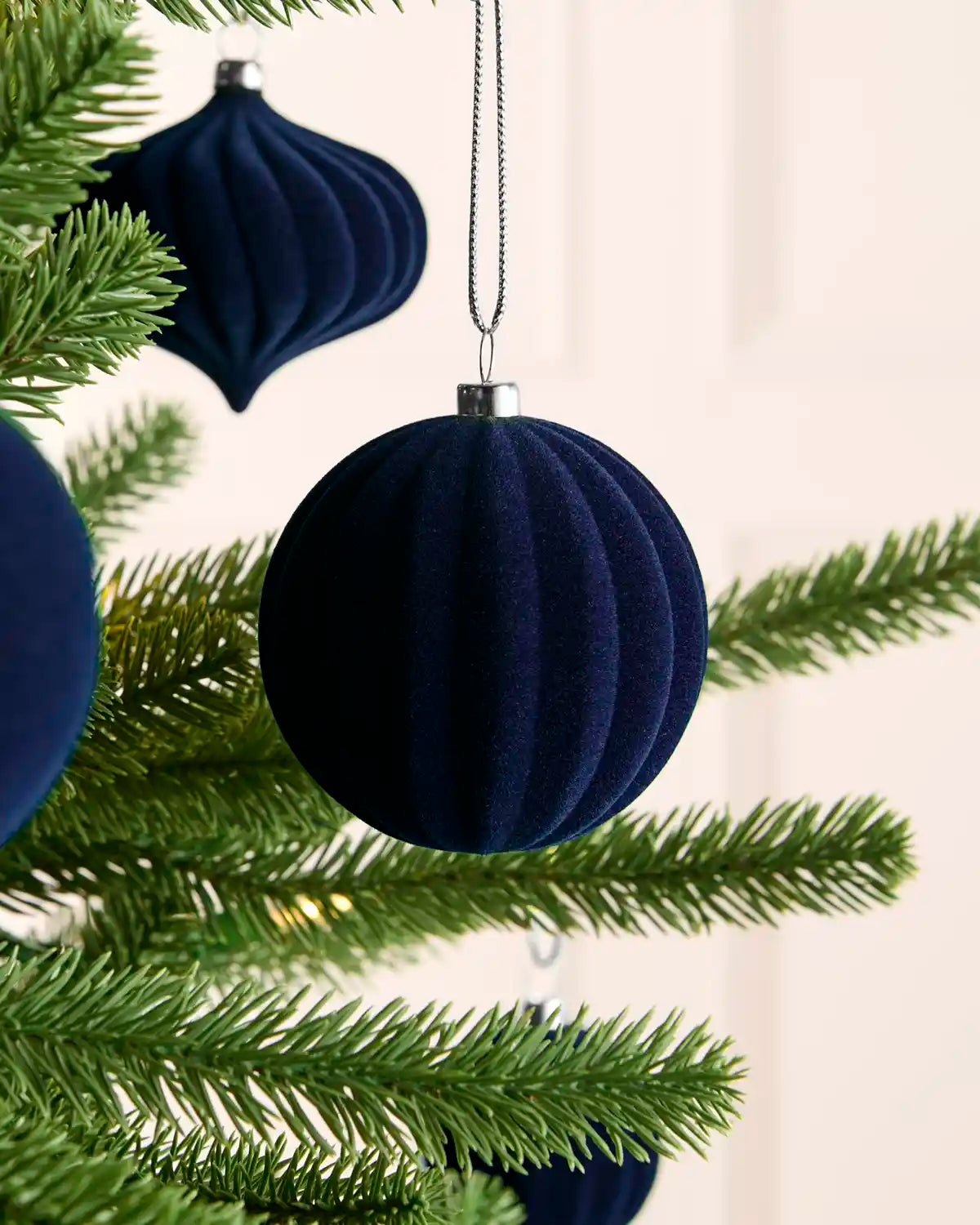 A dark navy blue ribbed velvet Christmas tree ornament hanging from a green pine tree branch against a soft off-white background.