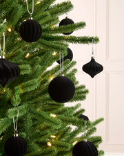 Black velvet Christmas tree ornaments hanging on a green artificial Christmas tree with warm white string lights, set against a light cream paneled wall.