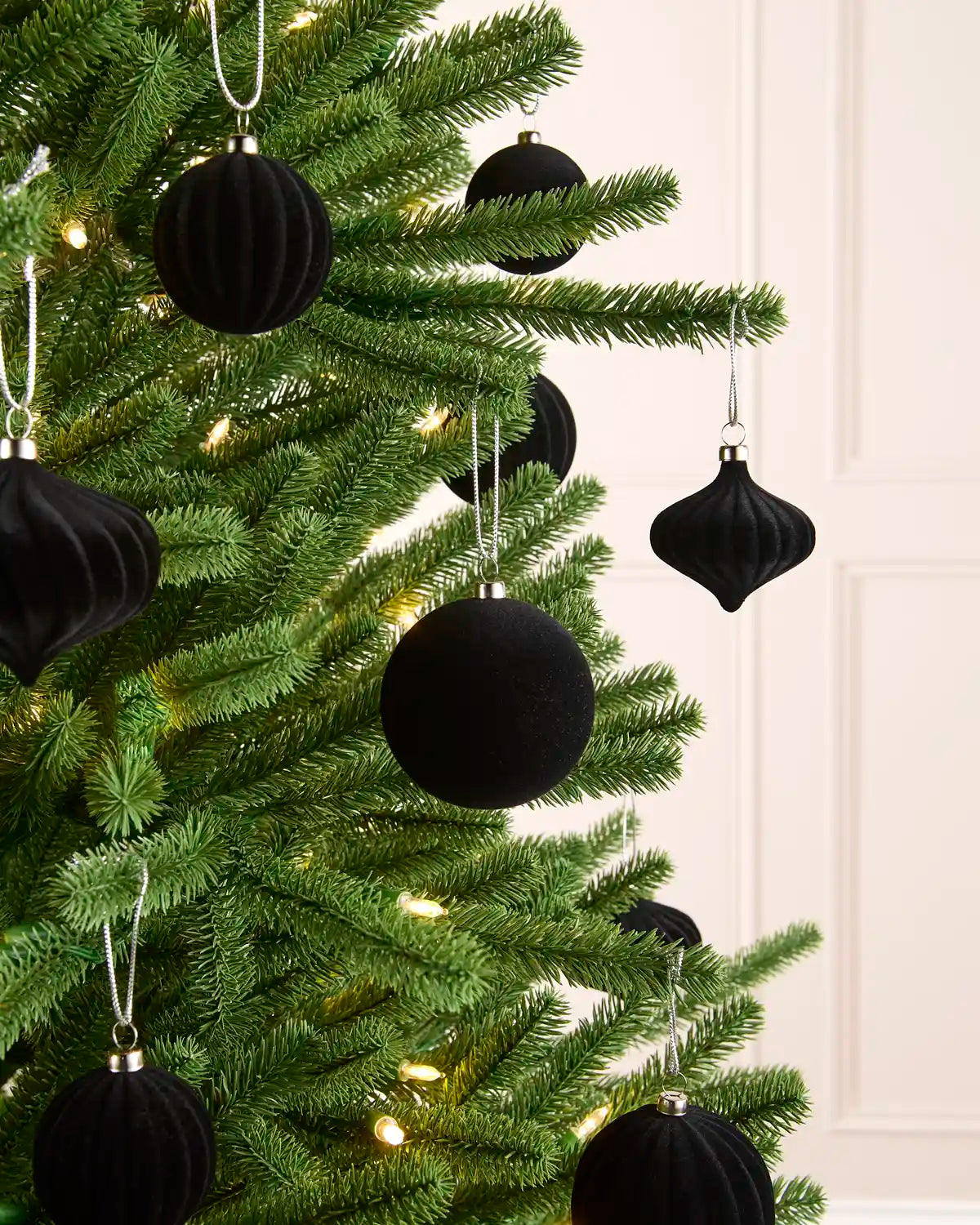 Black velvet Christmas tree ornaments hanging on a green artificial Christmas tree with warm white string lights, set against a light cream paneled wall.
