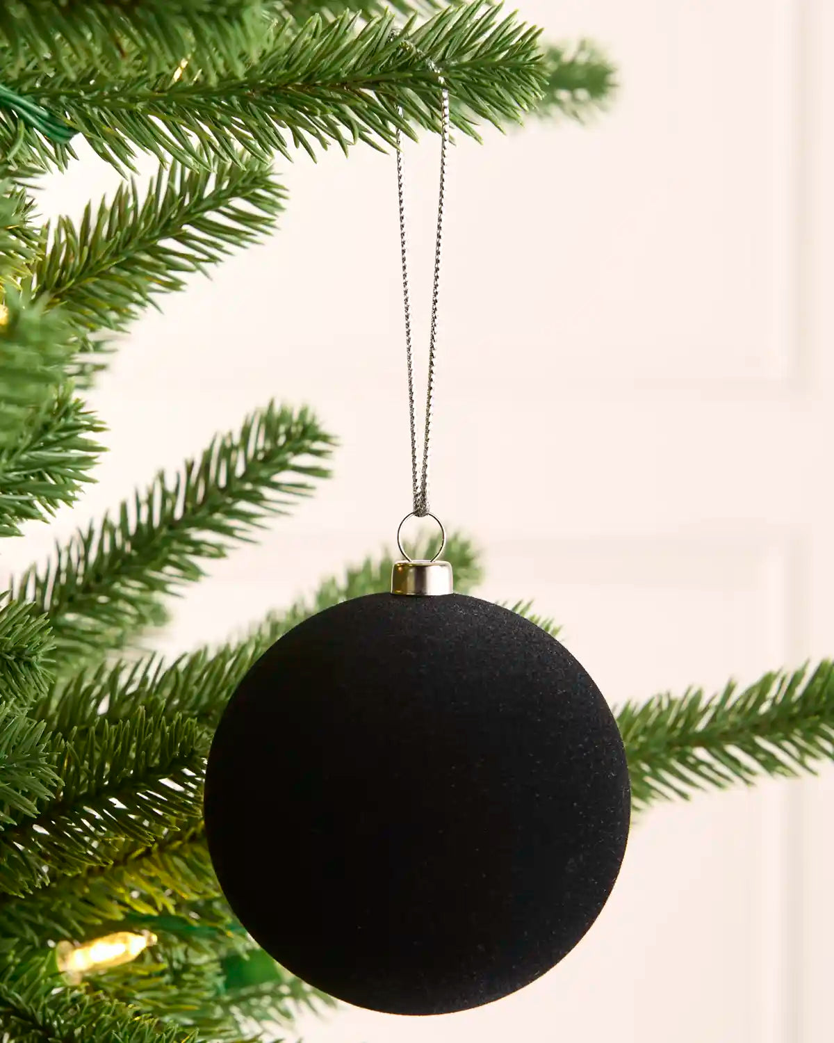 A velvet black spherical Christmas ornament hanging from a silver cap by a thin silver string, positioned in front of green pine tree branches with warm string lights against a soft off-white background.