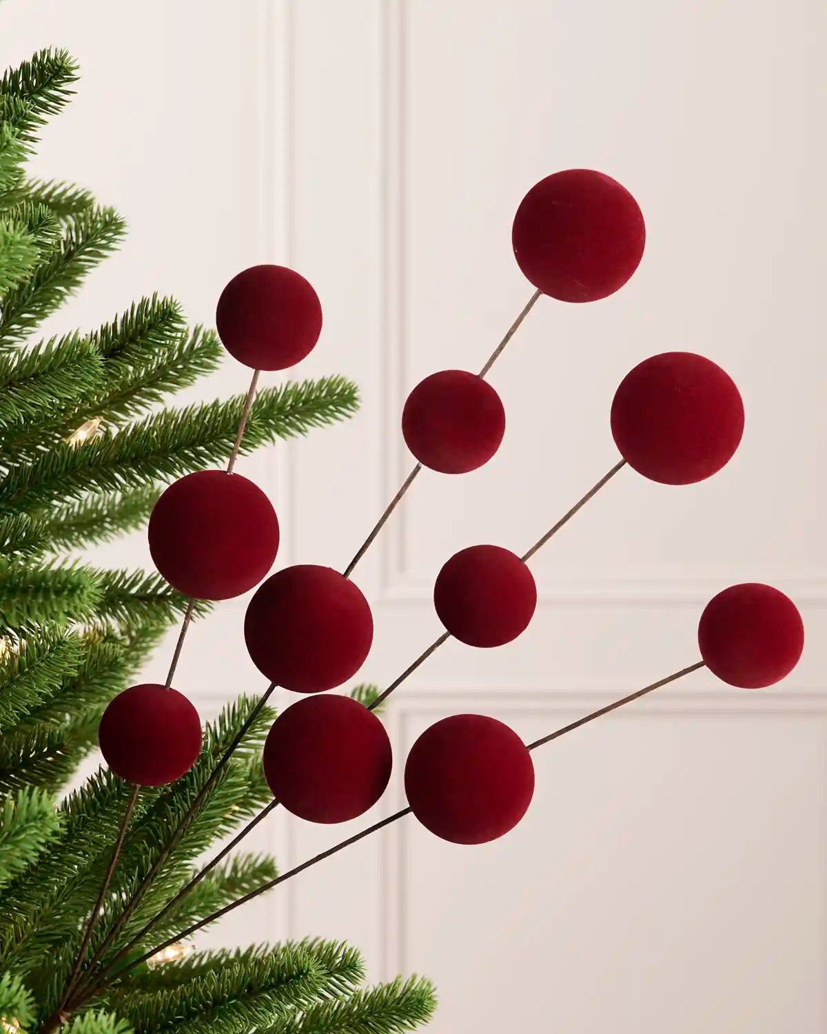 Burgundy velvet Christmas berry stems with multiple round burgundy balls on thin dark stems, styled on a green pine tree branch against a light cream panelled wall.