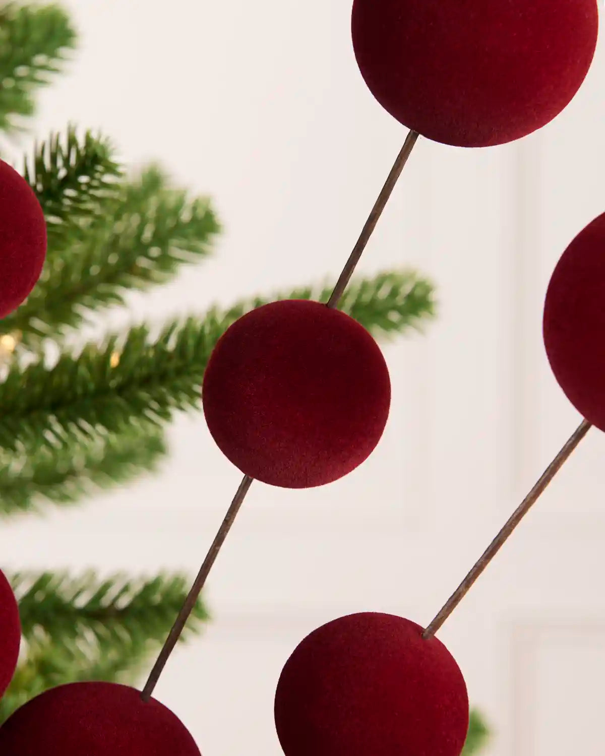Close up Burgundy velvet Christmas berry pick with multiple round burgundy balls on thin dark stems, styled on a green pine tree branch against a light cream panelled wall.