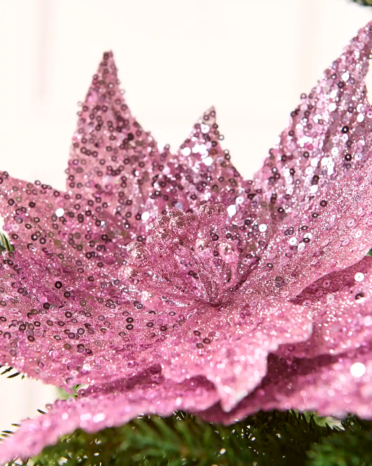 A close-up of a large pink sequined poinsettia decoration on green artificial pine branches against a soft off-white background.